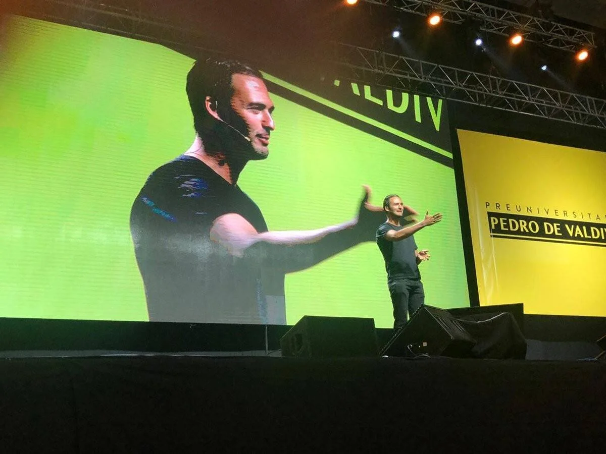 Un hombre con micrófono de diadema gesticula sobre un escenario frente a una gran pantalla que proyecta su imagen, en un evento del Preuniversitario Pedro de Valdivia.