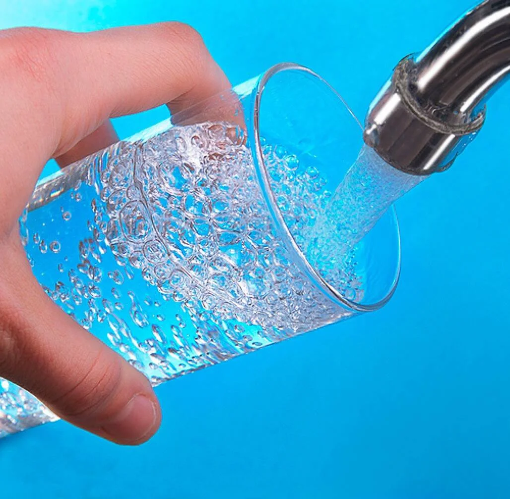 Una mano sostiene un vaso de vidrio bajo un grifo metálico del que cae agua con burbujas sobre fondo azul.