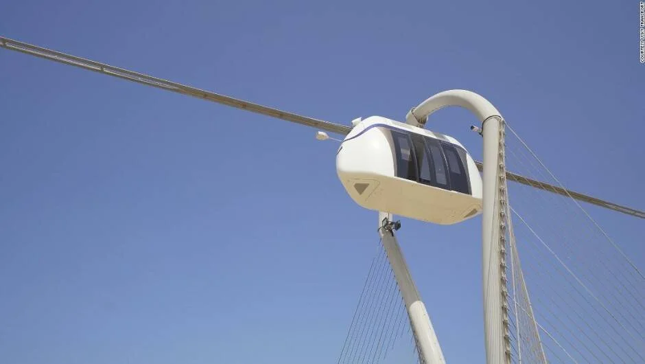 Pequeña cápsula de transporte autónomo blanca suspendida sobre un cable elevado contra un cielo azul despejado.