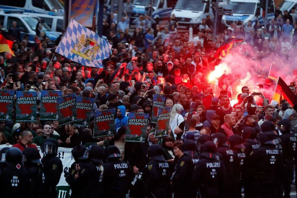 Multitud de manifestantes con carteles 'Asyflut Stoppen' y bengalas rojas, contenida por cordón policial en ciudad alemana.