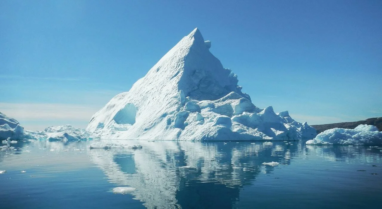 Gran iceberg de forma triangular con una caverna natural reflejado en aguas tranquilas bajo cielo despejado.