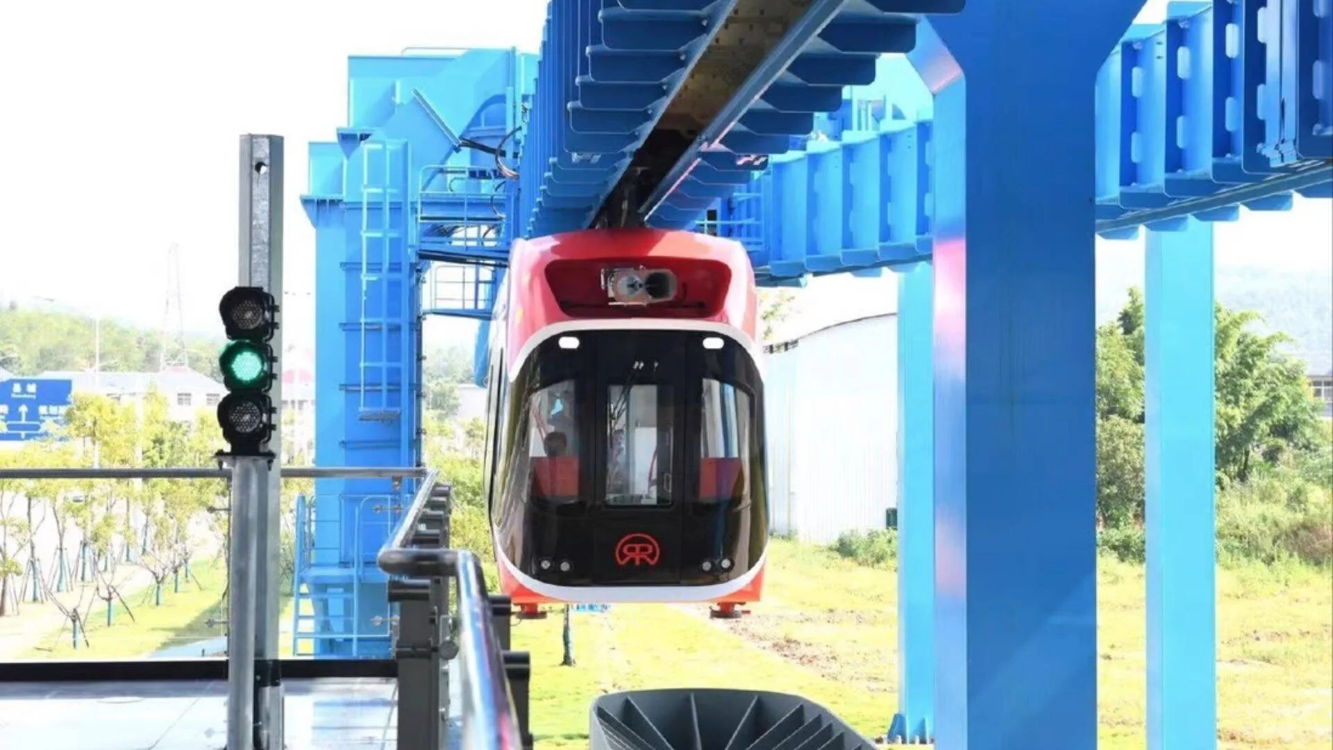 Vagón de tren suspendido colgante, de color rojo y blanco, circula bajo una estructura elevada de color azul junto a un semáforo en verde.