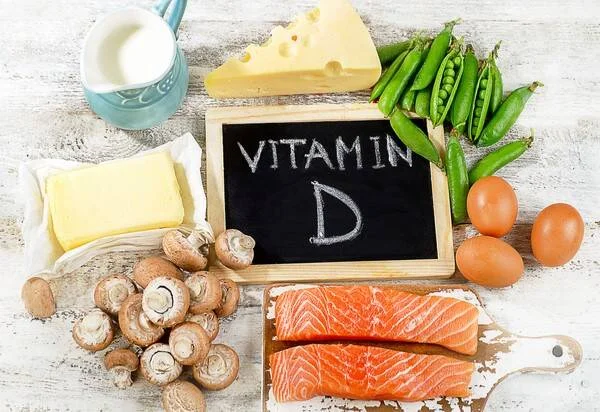 Variedad de alimentos fuente de vitamina D: salmón, huevos, hongos, queso, manteca, arvejas y leche junto a una pizarra con el texto 'Vitamin D'.