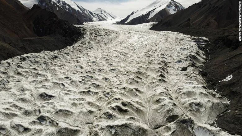 Vista aérea de un glaciar con superficie agrietada que desciende entre montañas rocosas bajo un cielo nublado.