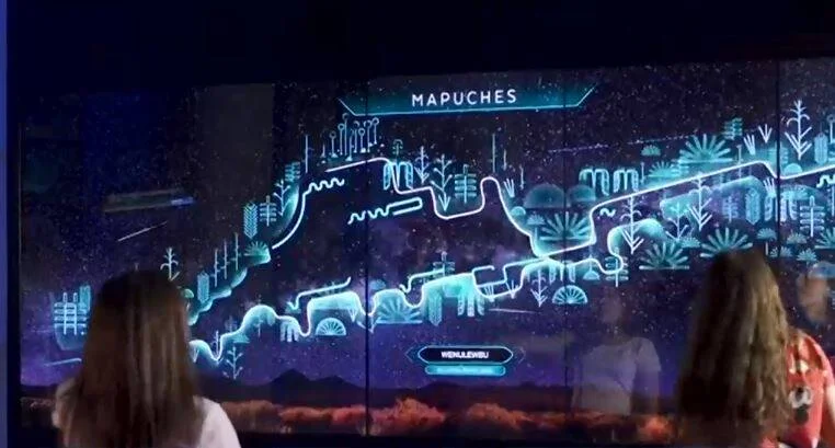 Dos visitantes observan una gran pantalla iluminada con animaciones digitales sobre la cultura mapuche.