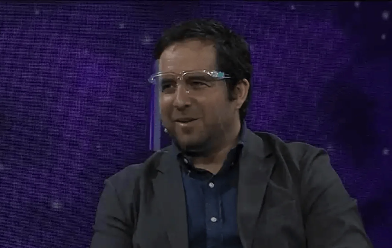 Hombre con protector facial transparente y saco gris sonríe en un estudio de televisión con fondo violeta.