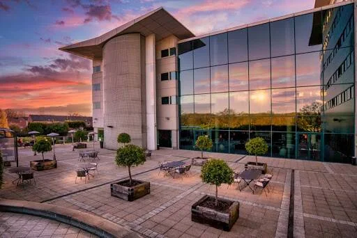 Patio exterior de un edificio moderno con fachada de vidrio, árboles en macetones y mesas al atardecer con cielo rojizo.