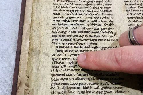 Una mano con anillo señala una línea de texto en un antiguo manuscrito medieval escrito en latín o francés antiguo.