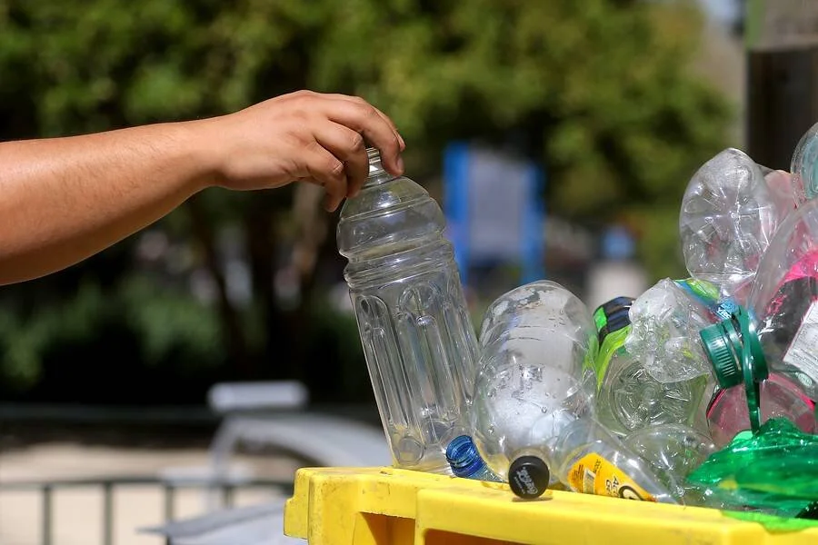 Una mano deposita una botella de plástico transparente en un contenedor amarillo lleno de envases para reciclar.