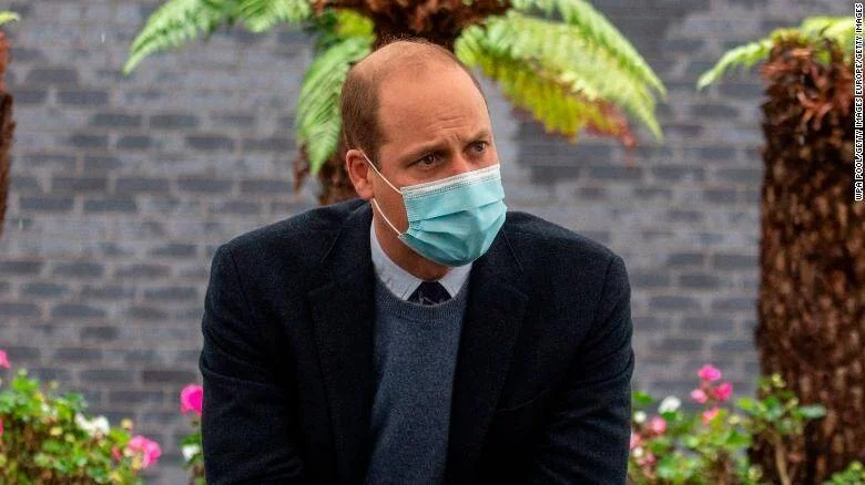 Hombre con mascarilla quirúrgica azul sentado en un jardín con flores rosadas y vegetación de fondo.