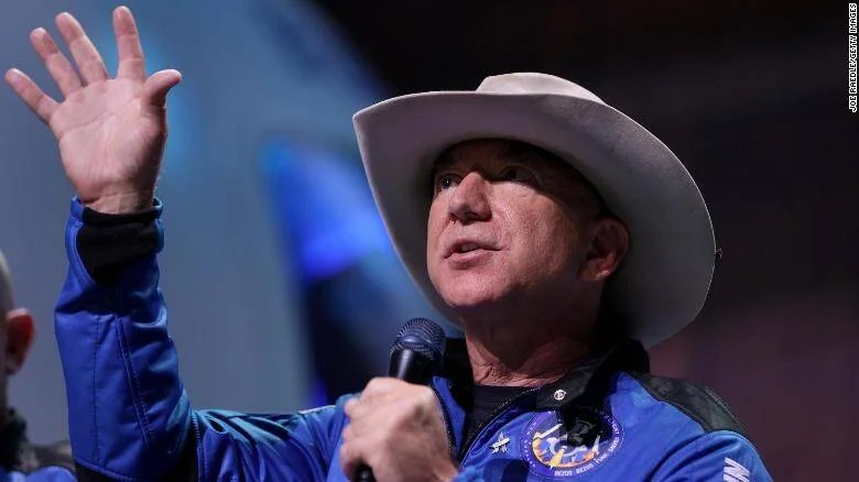 Hombre con sombrero vaquero blanco y traje azul de Blue Origin habla en un micrófono levantando la mano.