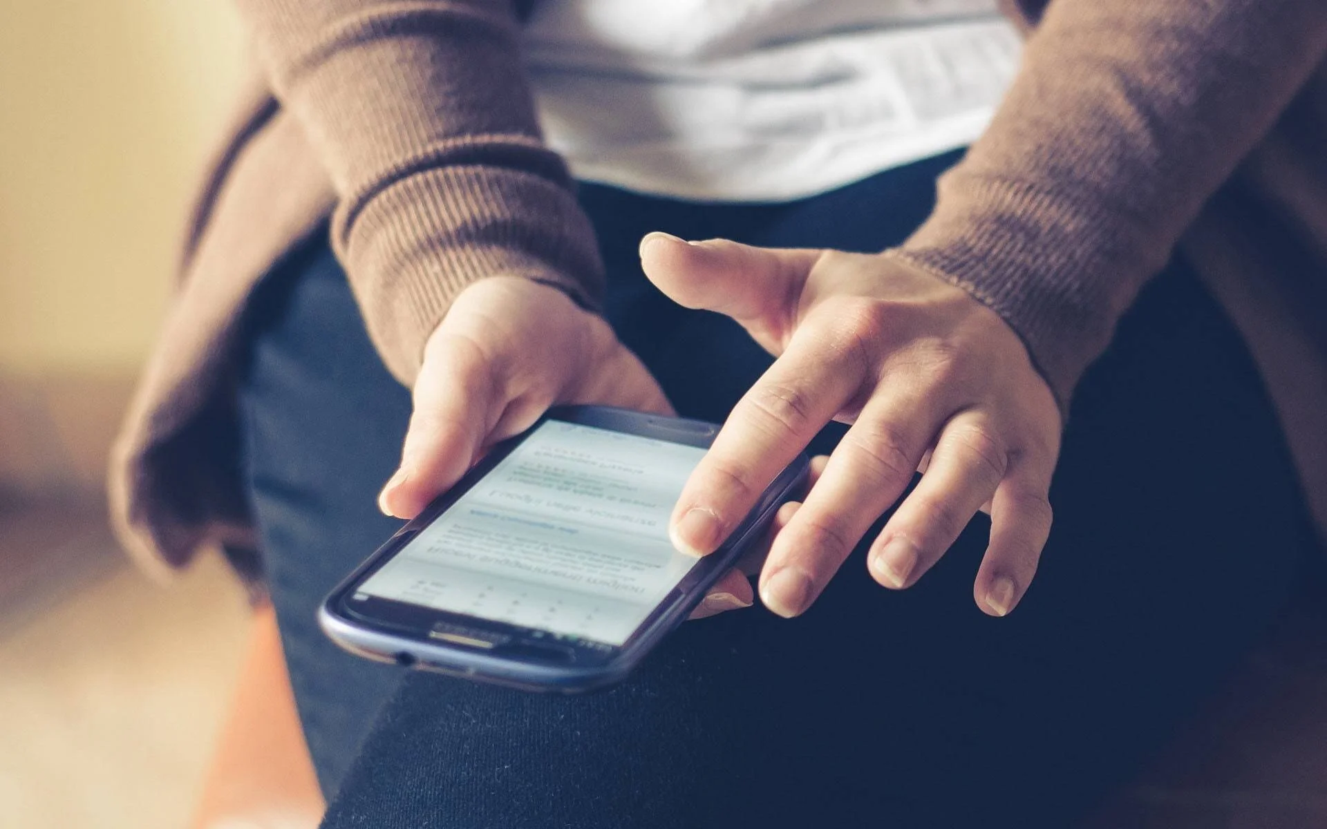 Manos de una persona sosteniendo un teléfono inteligente con texto en pantalla, apoyado sobre sus rodillas.