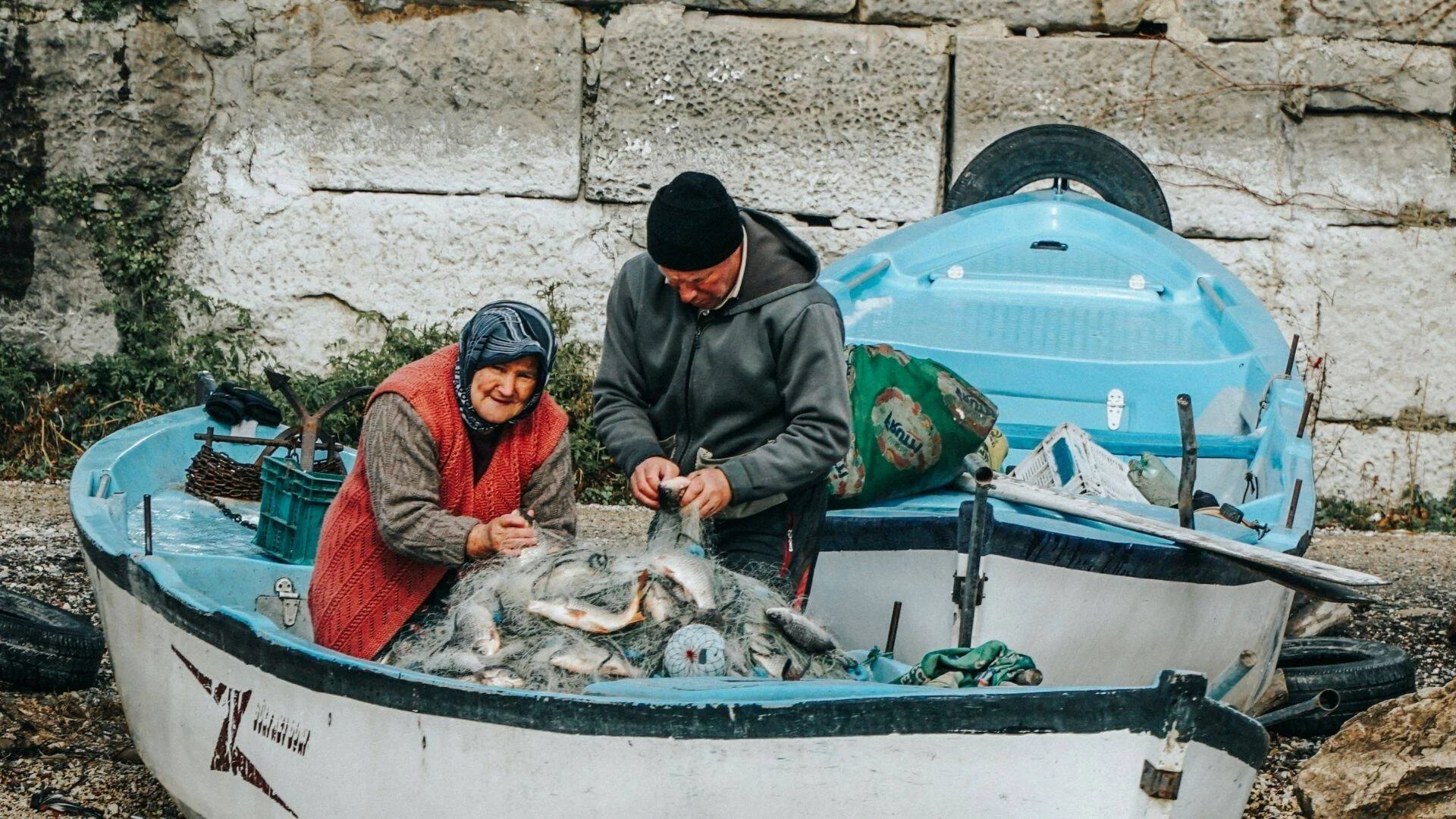 Una mujer mayor y un hombre joven clasifican peces atrapados en una red dentro de un bote de pesca azul y blanco en tierra.