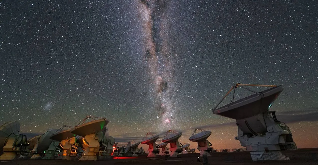 Conjunto de antenas de radiotelescopio apuntando al cielo nocturno con la Vía Láctea visible en el horizonte del desierto.