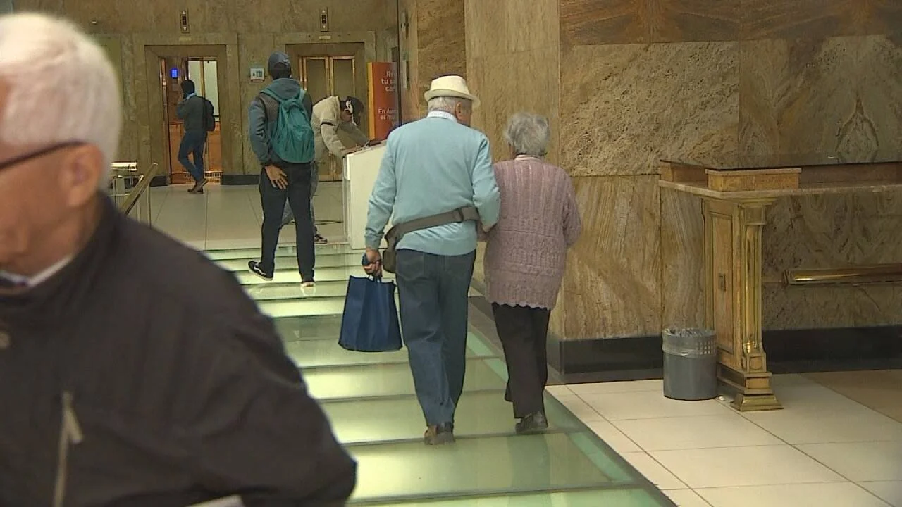 Pareja de personas mayores camina de espaldas por el interior de una entidad bancaria con columnas de mármol.