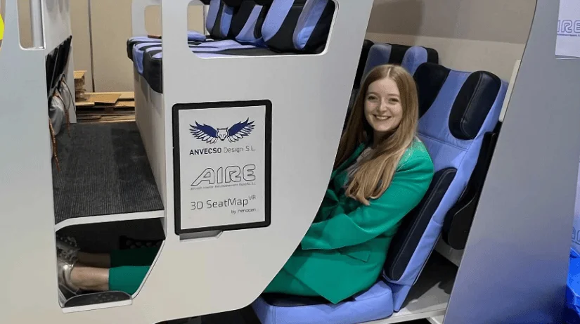 Una mujer sonriente prueba un asiento litera de dos niveles del concepto AIRE de Anvecso Design en una feria aeronáutica.