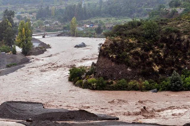 Río desbordado con aguas lodosas y turbulentas rodeando una formación rocosa durante una inundación.
