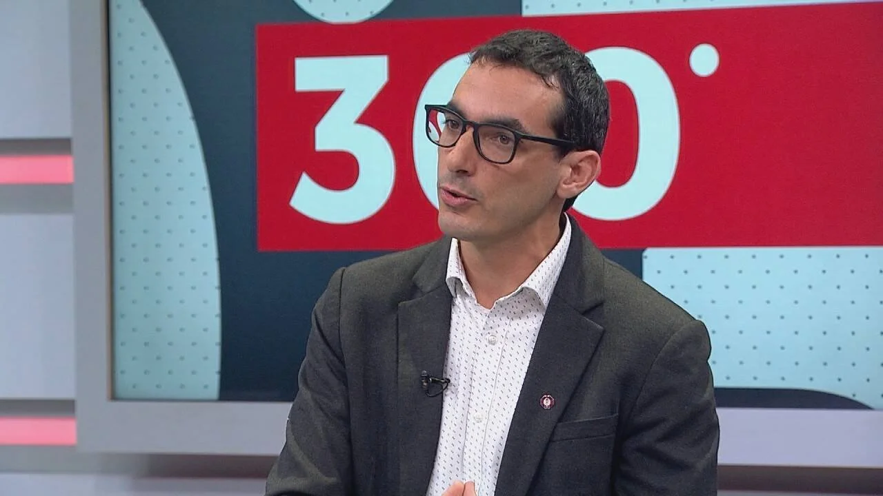 Hombre con anteojos y saco gris habla en un estudio de televisión con pantalla que muestra el logo '360°'.