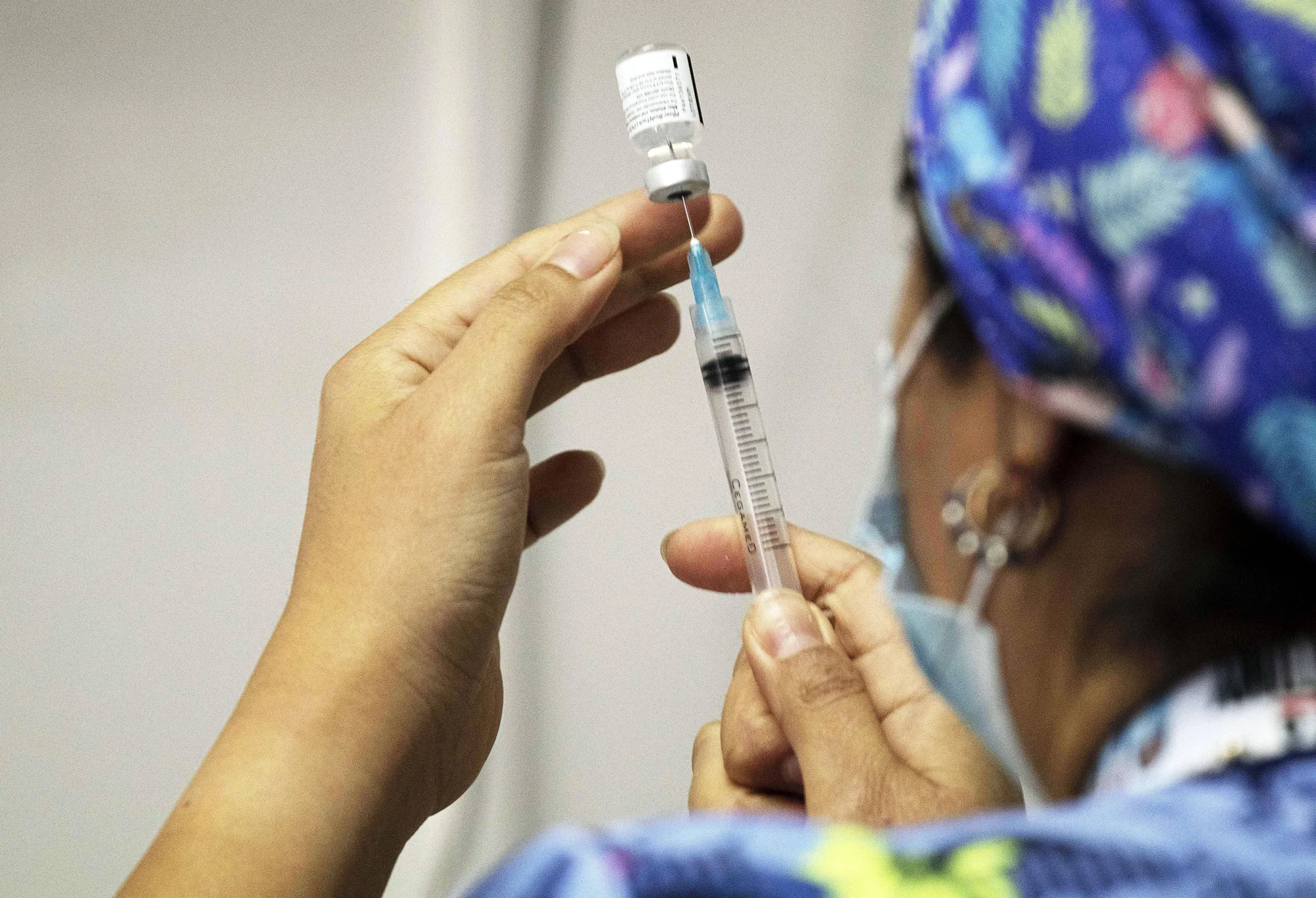 Una profesional de salud con mascarilla y gorro quirúrgico extrae una vacuna de un vial con una jeringa.