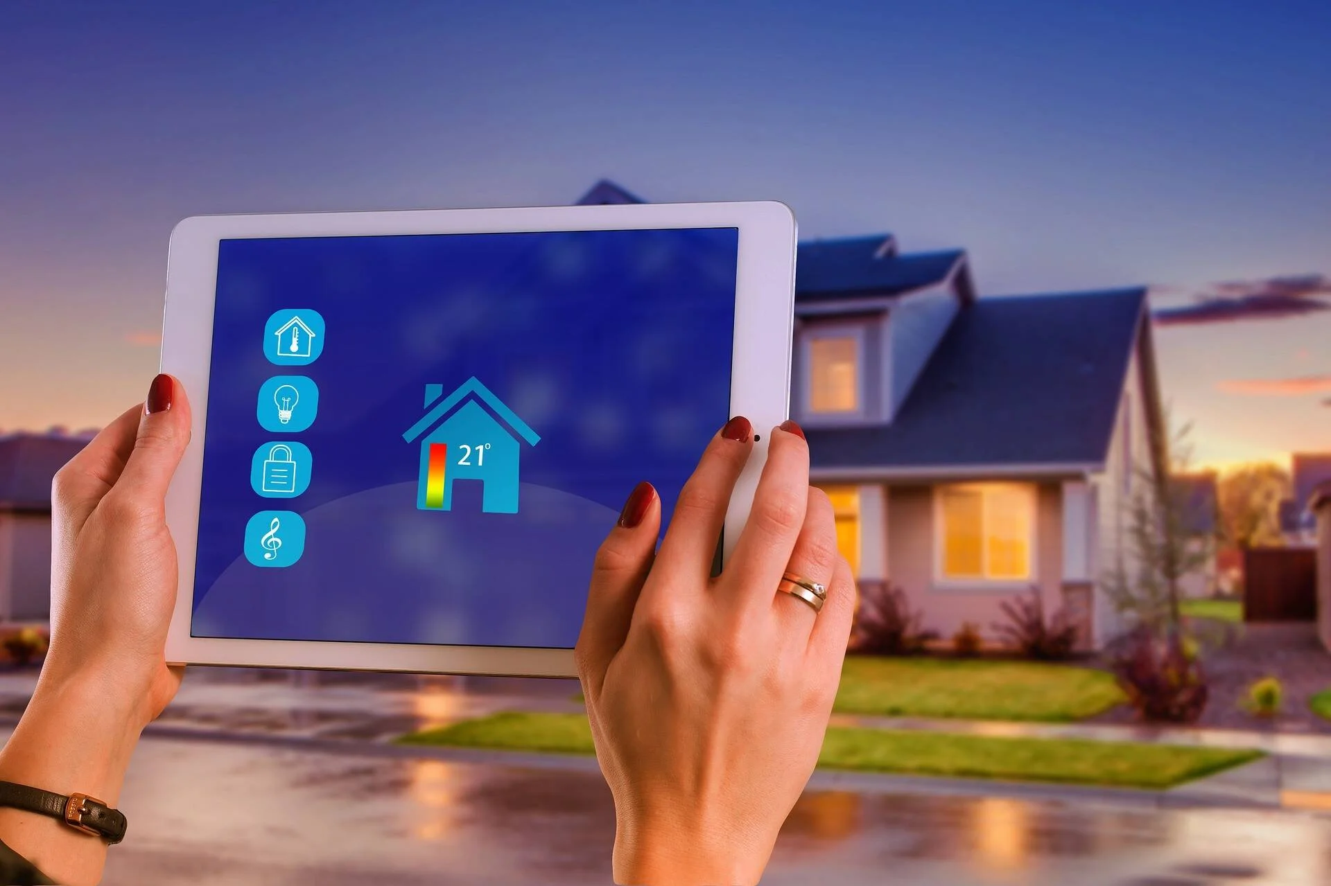 Manos sosteniendo una tablet con aplicación de domótica que muestra temperatura de 21° y controles del hogar, con una casa iluminada al fondo.