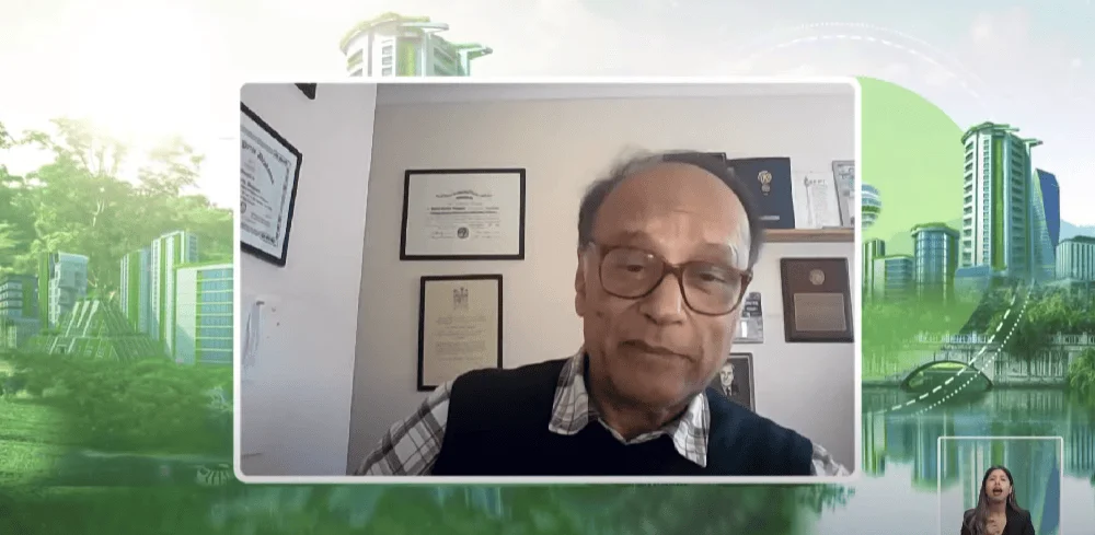 Un hombre mayor con lentes participa en una videollamada, con diplomas enmarcados en la pared detrás de él, en un evento sobre ciudades verdes.