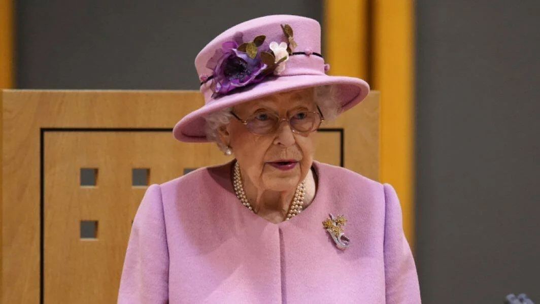 Mujer de edad avanzada vestida con traje y sombrero lila, collar de perlas y broche dorado, en un acto oficial.