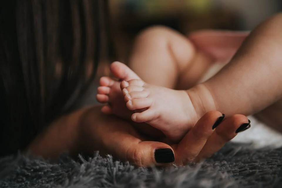 Una mano con uñas negras sostiene delicadamente los pequeños pies de un bebé sobre una alfombra gris.