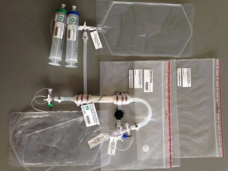 Conjunto de material médico que incluye jeringas, tubuladuras, llaves de paso y bolsas intravenosas etiquetadas sobre una superficie gris.