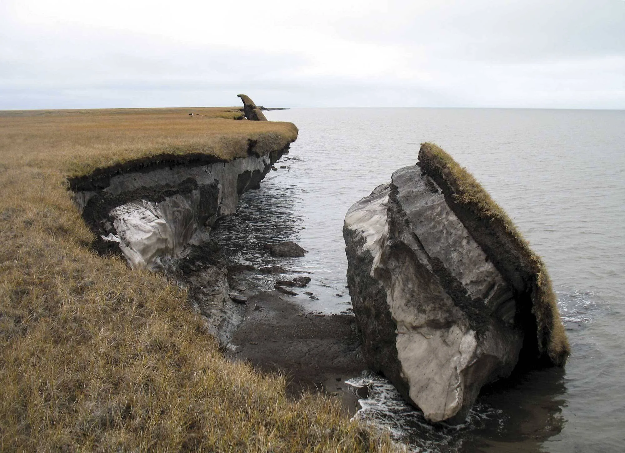 Acantilado de tundra con permafrost expuesto se derrumba al mar, mostrando bloques de hielo y suelo desprendidos.