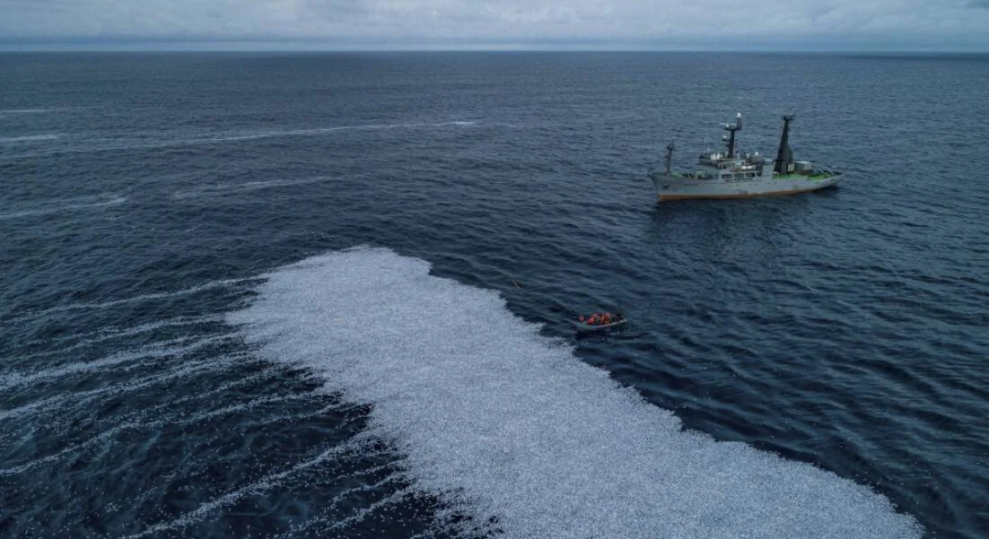 Vista aérea de un buque pesquero en alta mar junto a una embarcación menor, con una gran mancha blanquecina flotando en la superficie del océano.