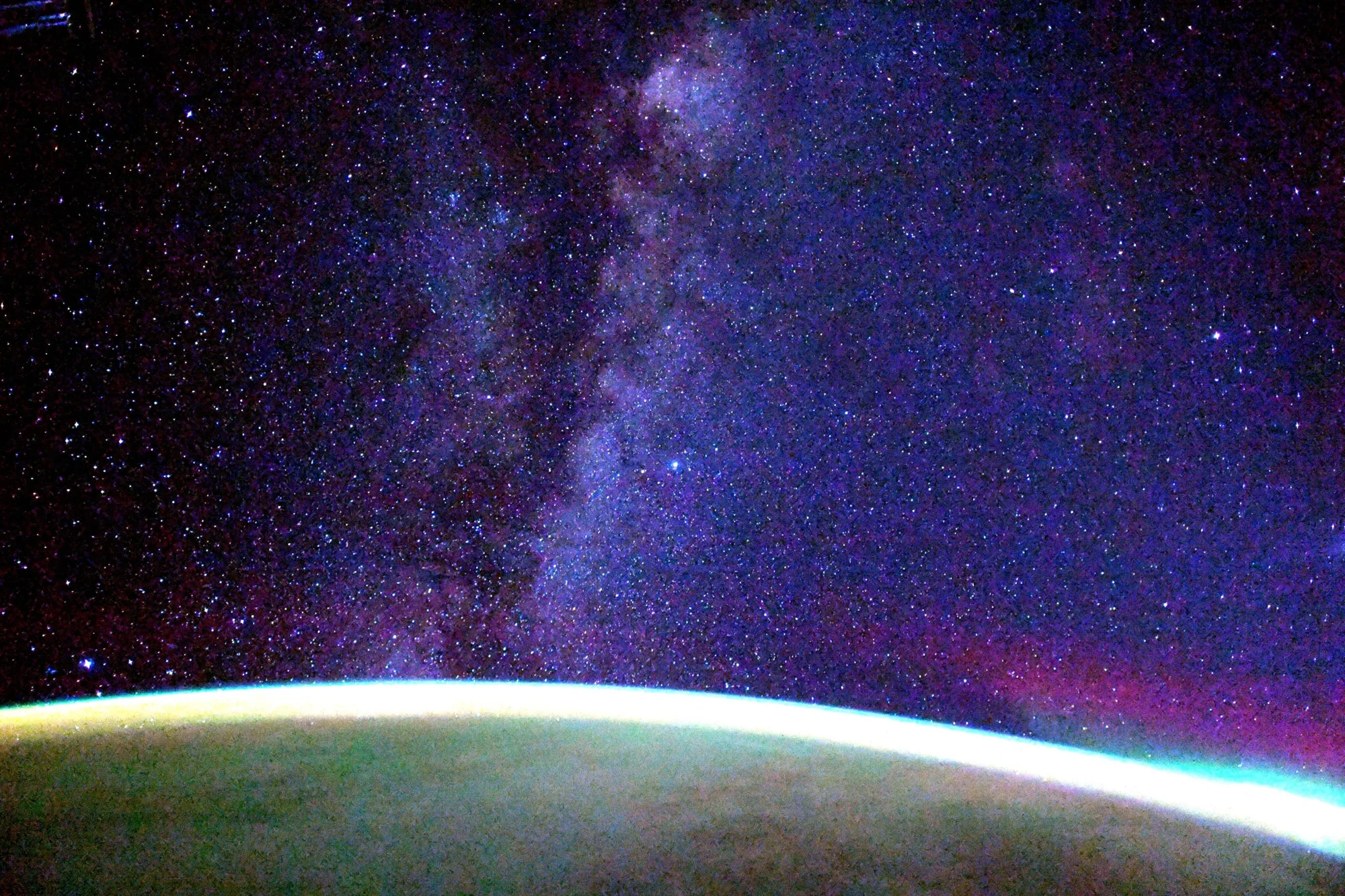La Vía Láctea con miles de estrellas y nebulosas violáceas se extiende sobre el horizonte curvo iluminado de la Tierra visto desde el espacio.