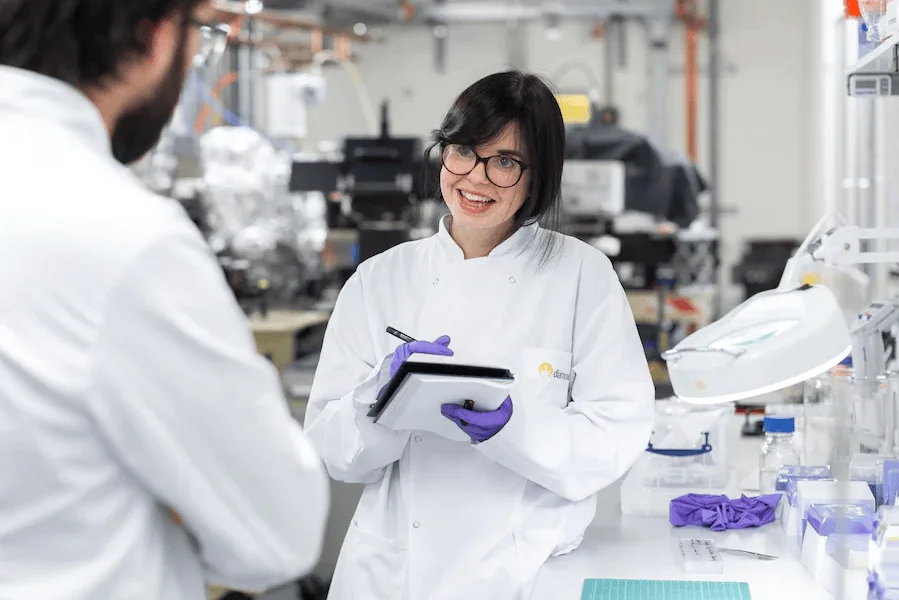 Investigadora con bata blanca y guantes morados toma notas en una tablet mientras conversa con un colega en un laboratorio.