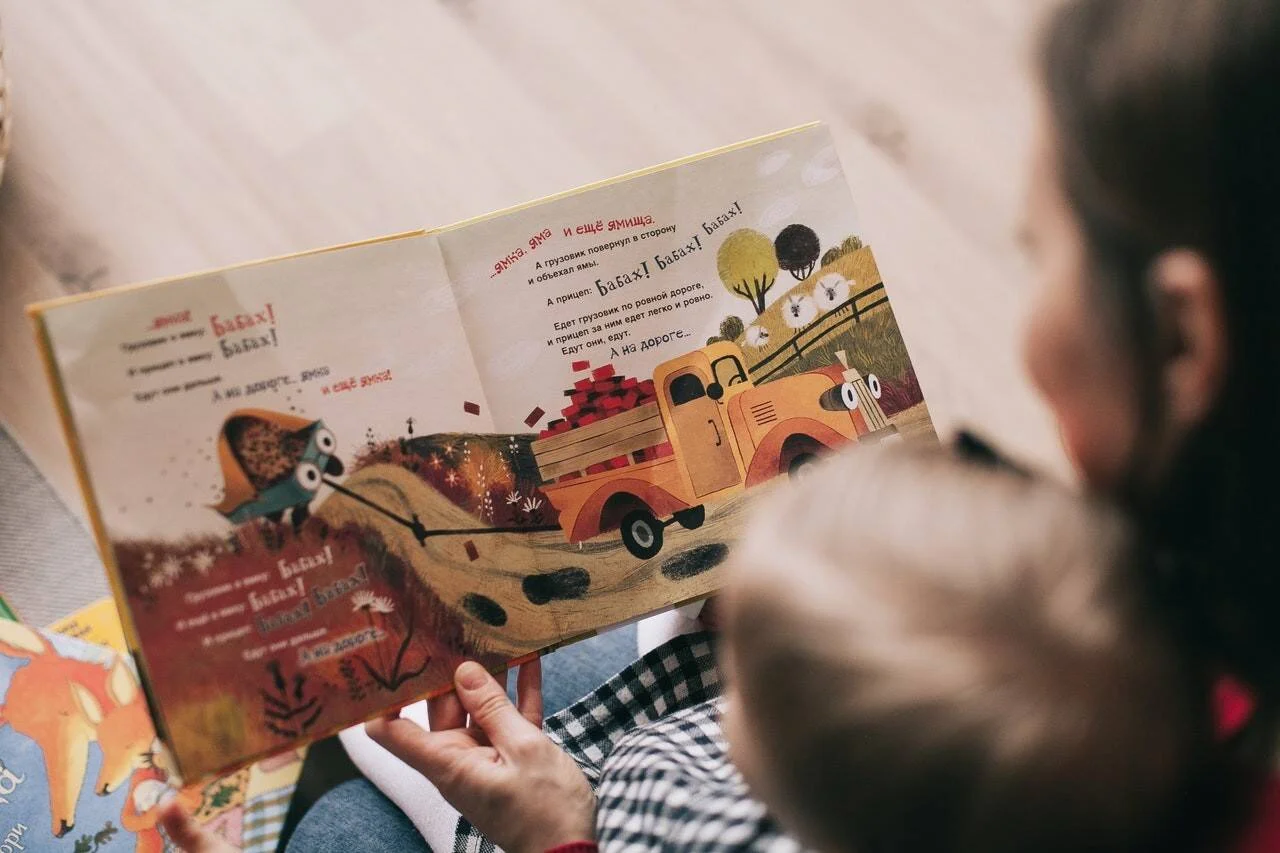 Un adulto sostiene un libro ilustrado infantil en ruso con dibujos de un camión, mientras un niño lo observa.