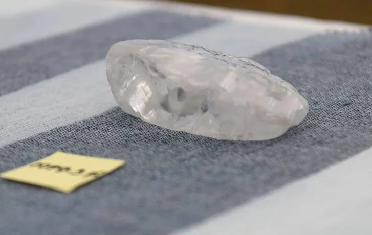 Gran diamante en bruto de color blanco translúcido sobre una tela gris, junto a una etiqueta amarilla con inscripción.