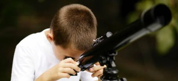 Un niño con camiseta blanca observa a través de un telescopio negro en un entorno natural.