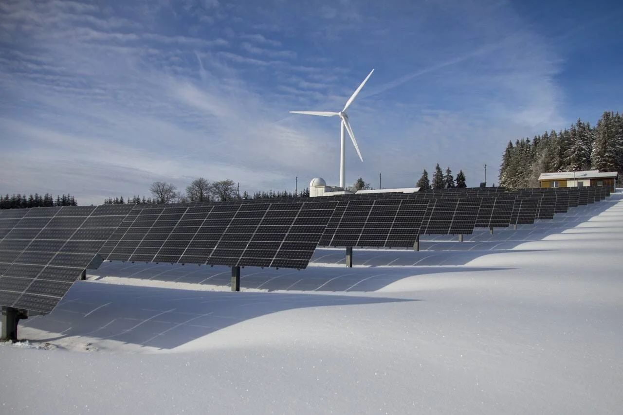 Granja de paneles solares cubierta de nieve con un aerogenerador y bosque de coníferas al fondo en un día despejado.