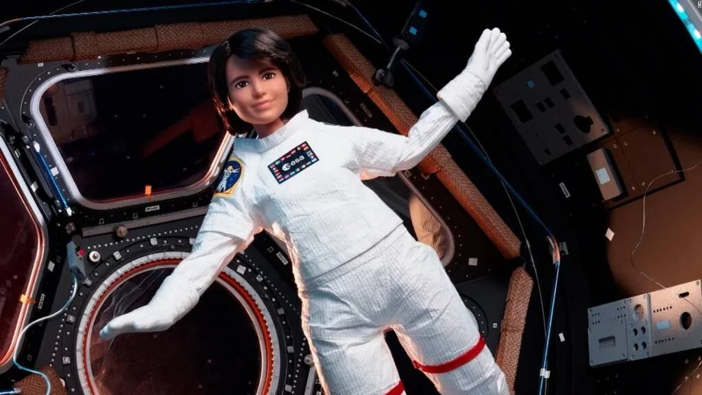 Muñeca Barbie vestida con traje espacial blanco de la ESA, flotando dentro de una estación espacial en microgravedad.