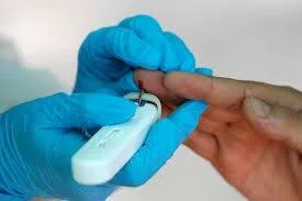 Profesional con guantes azules realiza punción en el dedo de un paciente con un dispositivo de prueba rápida.