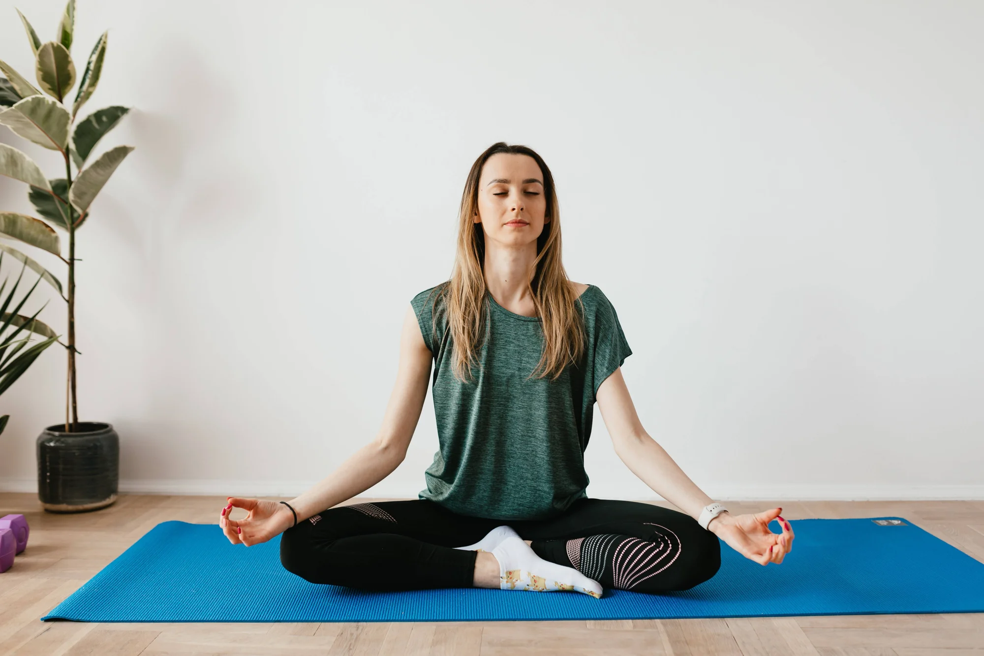 Mujer joven sentada en postura de loto sobre una esterilla azul, meditando con los ojos cerrados en un espacio interior.