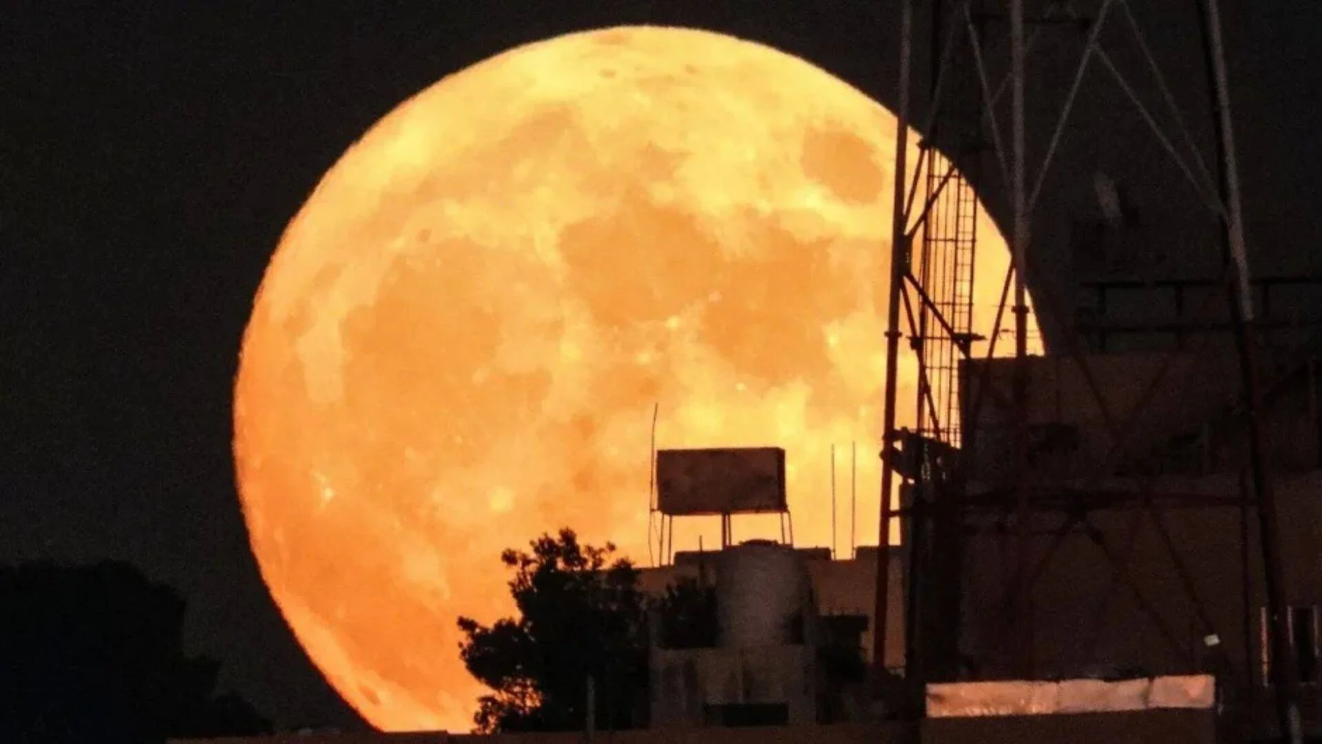 Luna llena de gran tamaño y tonalidad naranja intensa se eleva detrás de edificios y estructuras metálicas en la noche.