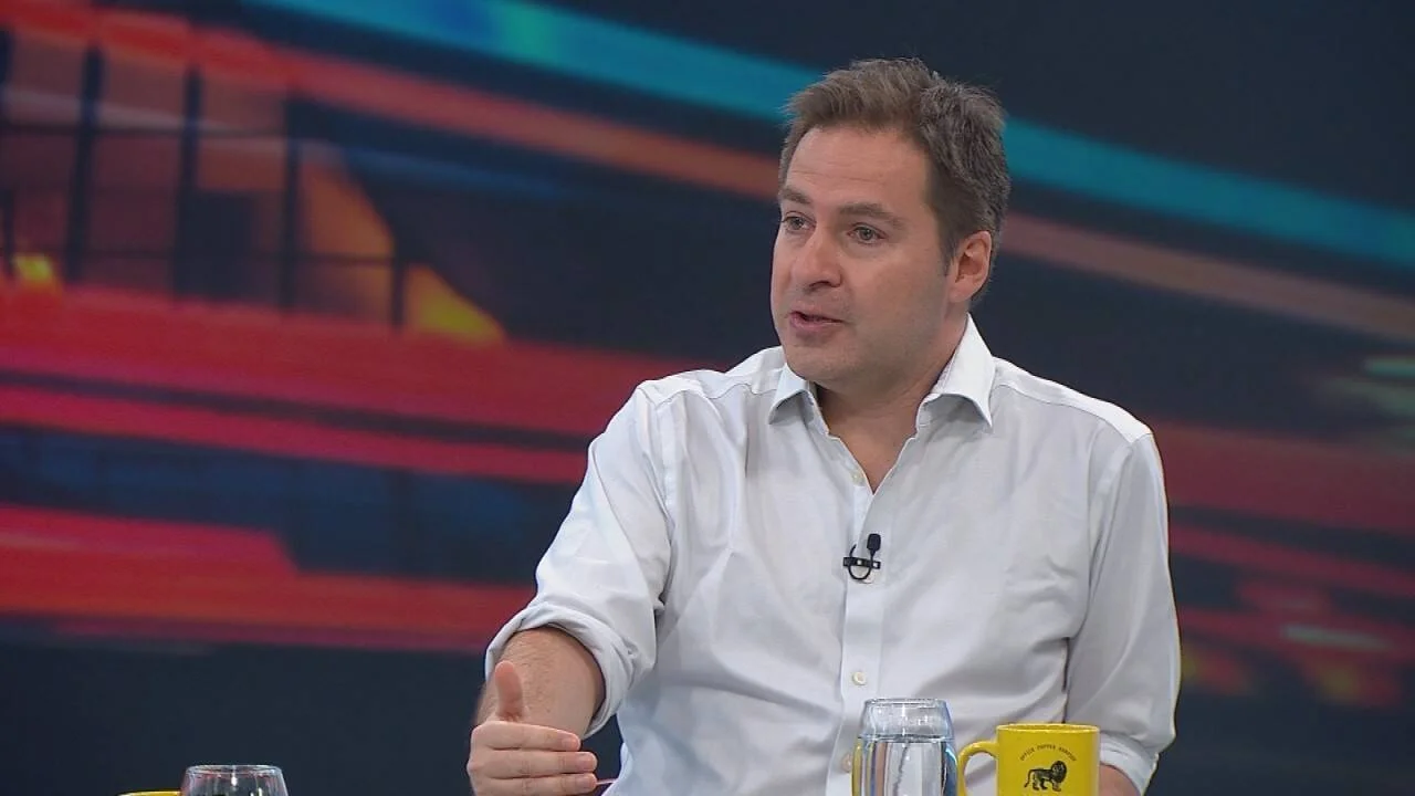Hombre de camisa blanca habla con micrófono de solapa en un estudio de televisión con fondo colorido.