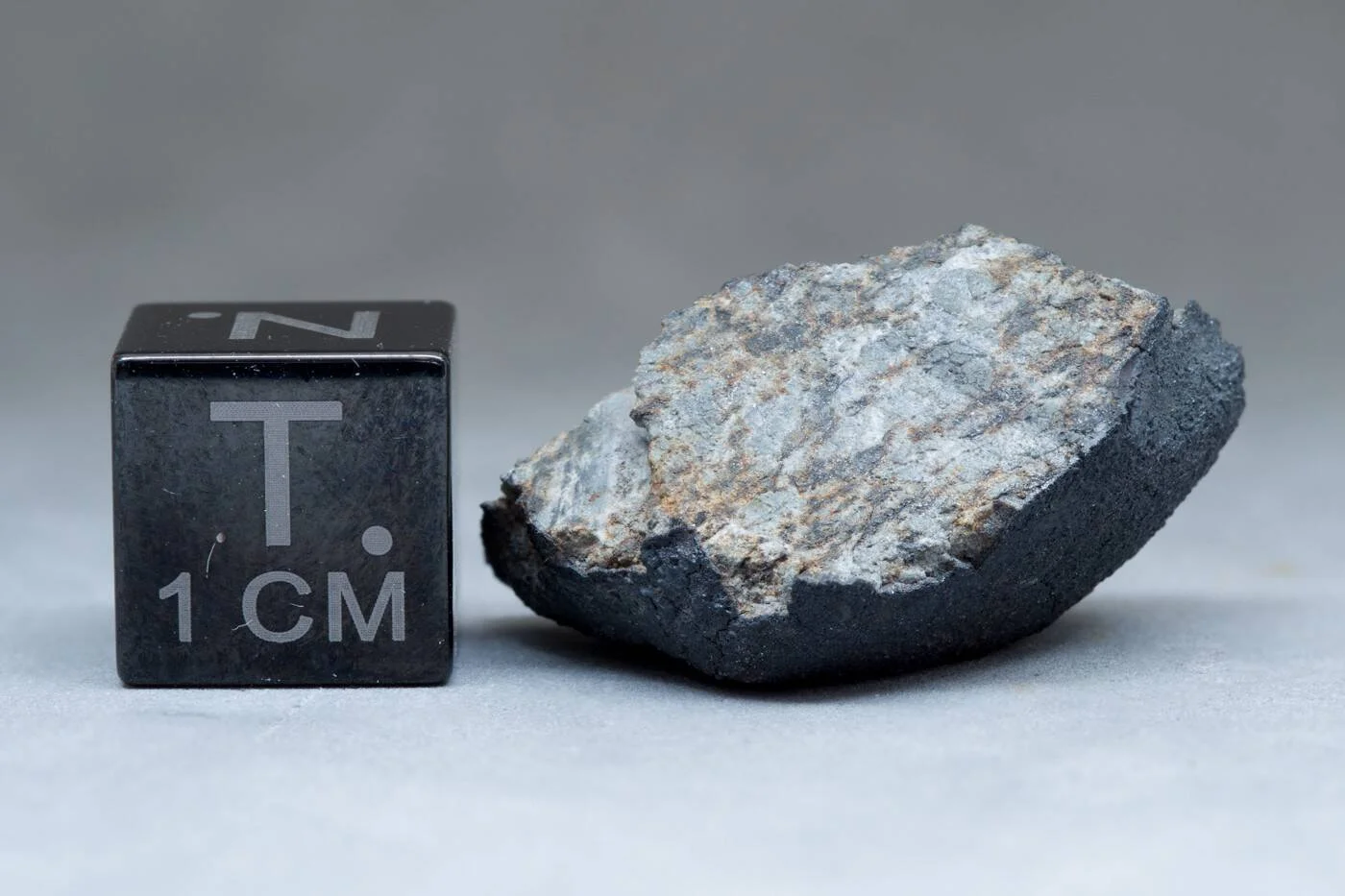 Fragmento de meteorito con corteza de fusión oscura e interior gris claro, junto a un cubo de referencia de 1 cm.