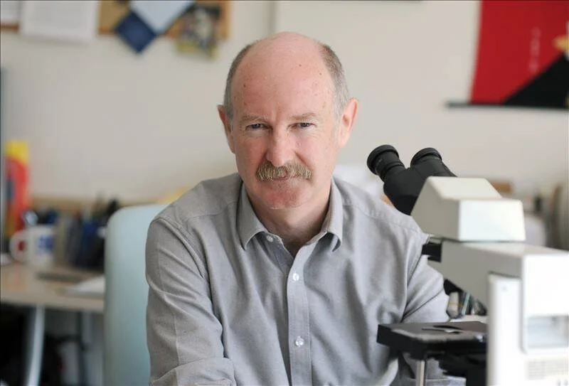Hombre de mediana edad con bigote sentado junto a un microscopio en un entorno de laboratorio u oficina científica.