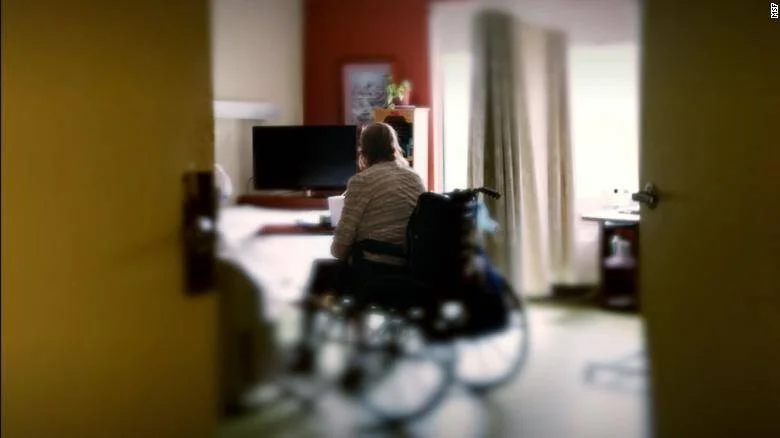 Persona en silla de ruedas de espaldas, sola en una habitación frente a un televisor, vista desde el umbral de una puerta.