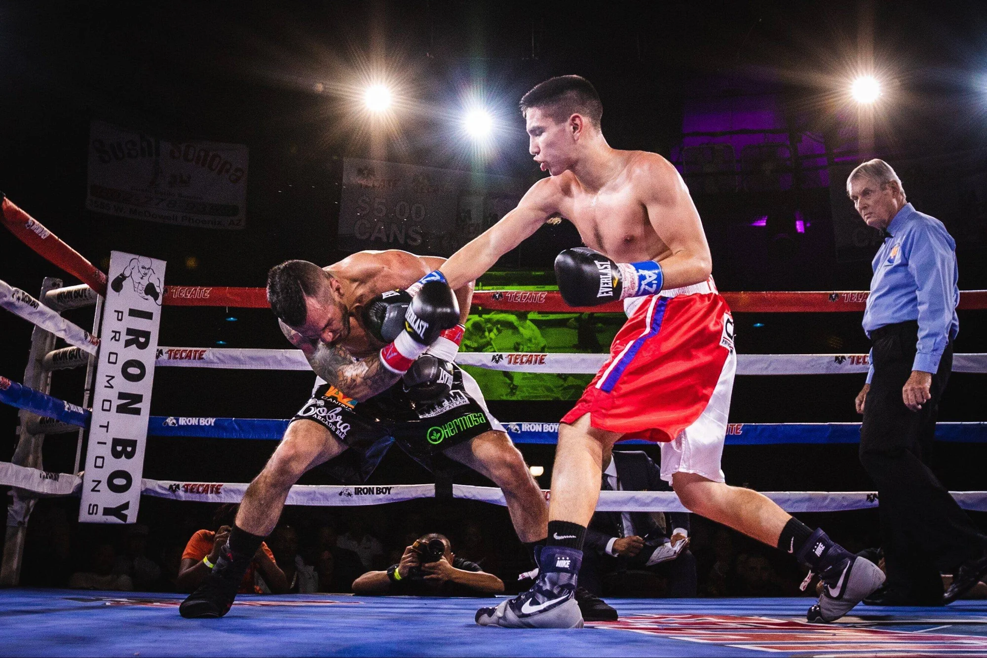 Boxeador con shorts rojos conecta un contundente golpe a su rival durante una pelea nocturna de Iron Boy Promotions.