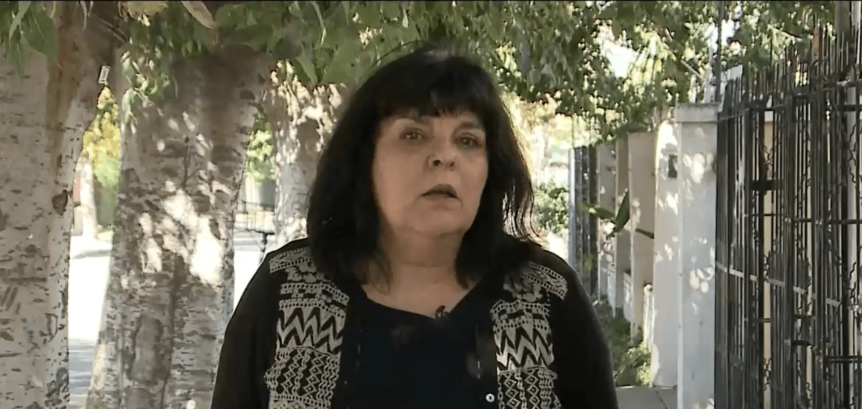 Mujer de cabello negro con blusa estampada habla frente a cámara en una calle arbolada.