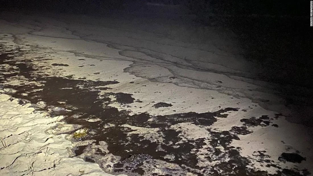 Manchas oscuras de petróleo mezcladas con espuma blanca en la orilla de una playa, fotografiadas de noche con linterna.
