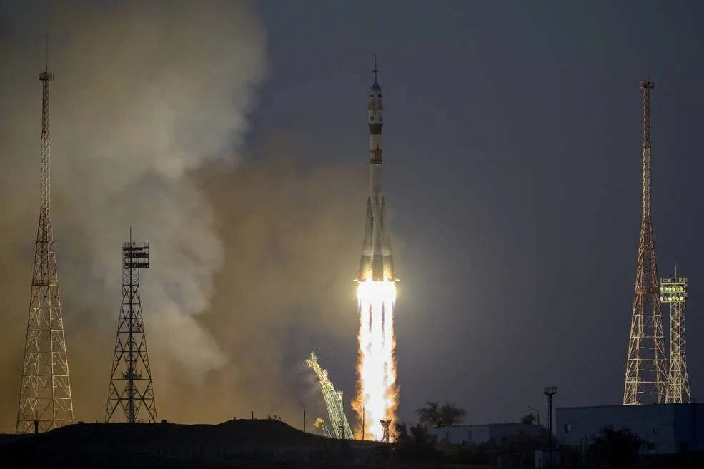 Cohete Soyuz despega entre llamas y columnas de humo desde una plataforma de lanzamiento al anochecer.