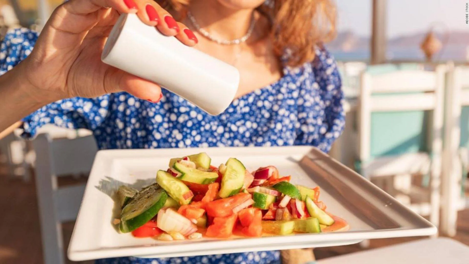 Una mujer condimenta con un salero una ensalada de tomate, pepino y cebolla roja en un plato blanco.