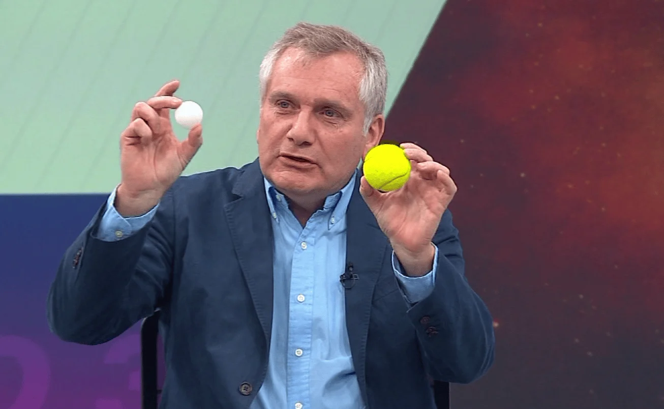 Hombre con saco azul sostiene una pelota blanca de ping pong y una pelota amarilla de tenis frente a cámara en un estudio de televisión.